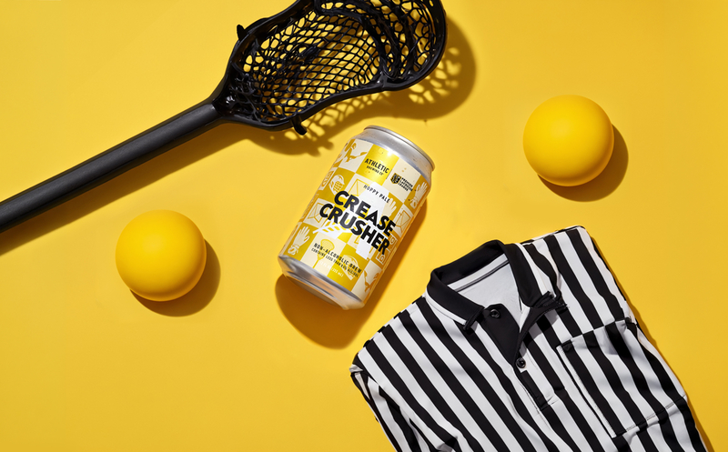 Lacrosse gear on a bright yellow background: stick head, two yellow balls, a striped polo shirt, and a can labeled 'Grease Crusher'.