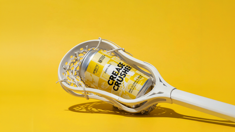Lacrosse stick head holding a yellow can labeled 'Crease Crusher' on a bright yellow background from Athletic Brewing.