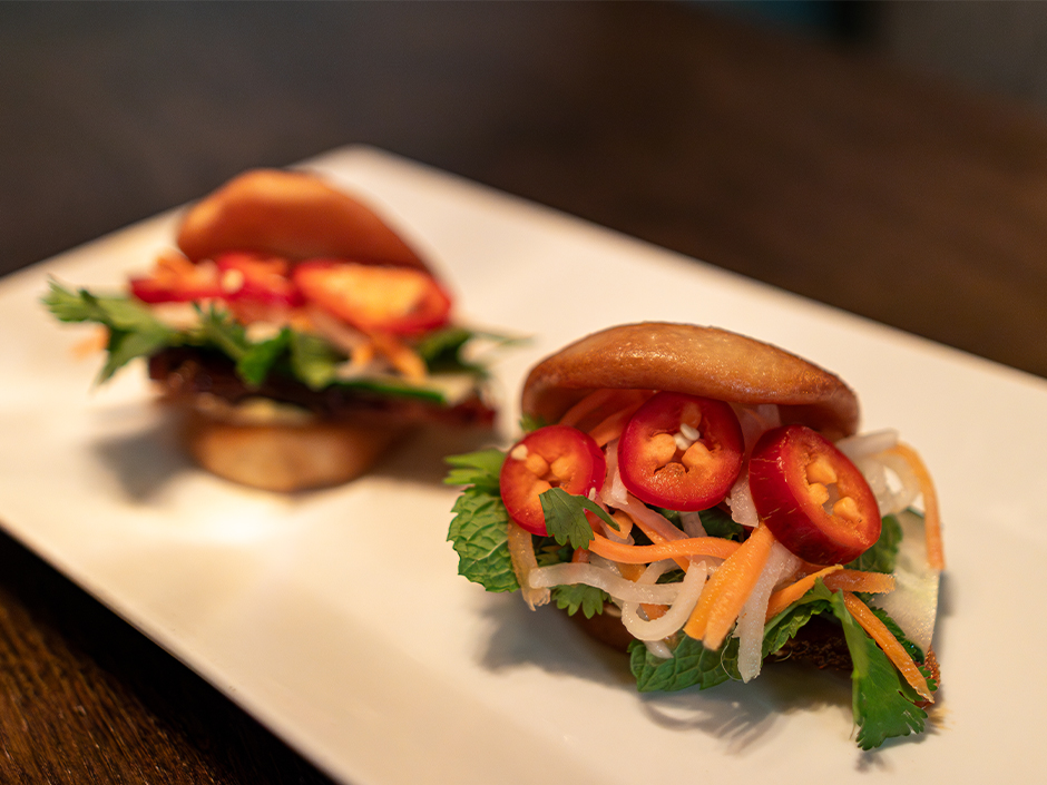 Two mini bao buns filled with fresh herbs, shredded vegetables, and sliced red chilies on a white plate.