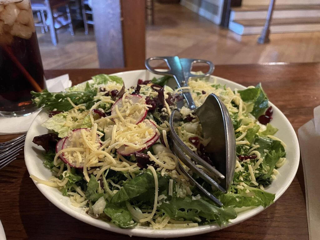 Bowl of mixed green salad with shredded cheese, radish slices, and a metal serving utensil on a wooden table; a drink with a straw is visible on the left