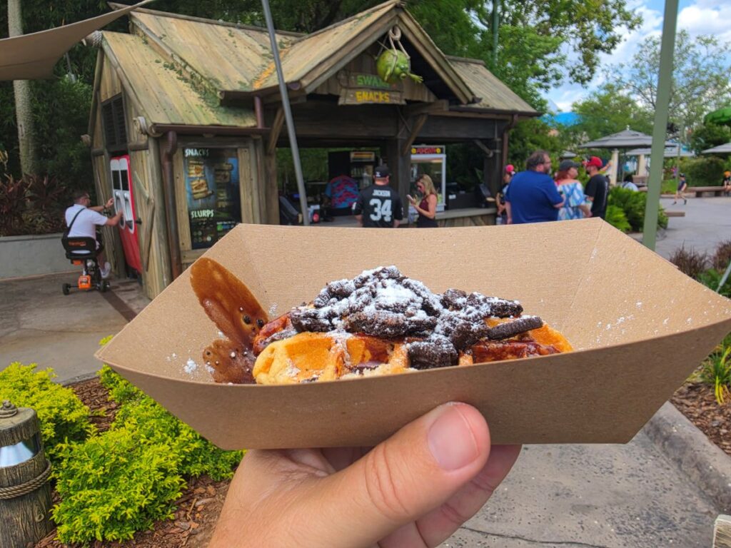 Person holding a brown paper boat filled with powdered-sugar dusted fried pastries topped with chocolate chunks in an outdoor snack stand area, which is Donkey's Triple Chocolate Waffle