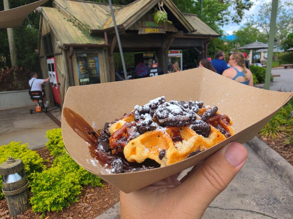 Hand holding a boat-shaped cardboard tray with Donkey’s Triple Chocolate Waffle dessert topped with chocolate pieces and powdered sugar outdoors near a rustic snack shack.