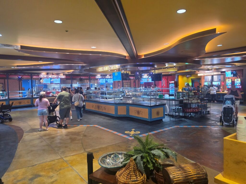 Indoor food court with multiple counter stalls and yellow accents, customers in line and strollers nearby.