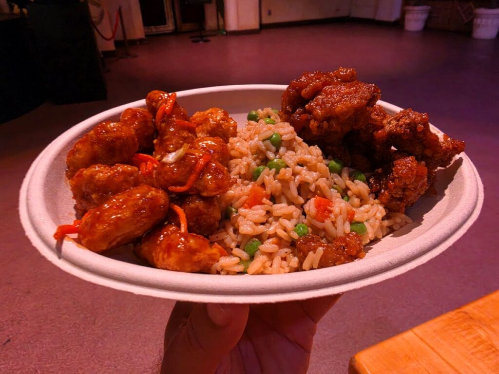 Plate of glazed fried chicken pieces with peas and diced carrots mixed into fried rice on a white disposable bowl surface visible in a dim, pink-lit setting.