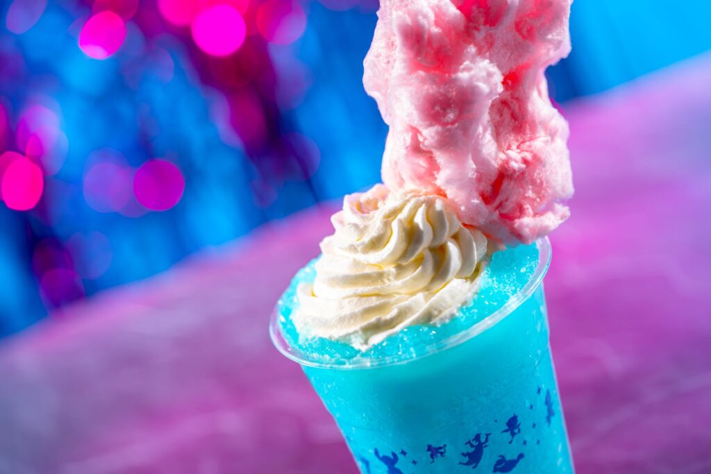 Blue milkshake in a clear cup topped with whipped cream and pink cotton candy, set against a colorful blue and pink bokeh background.