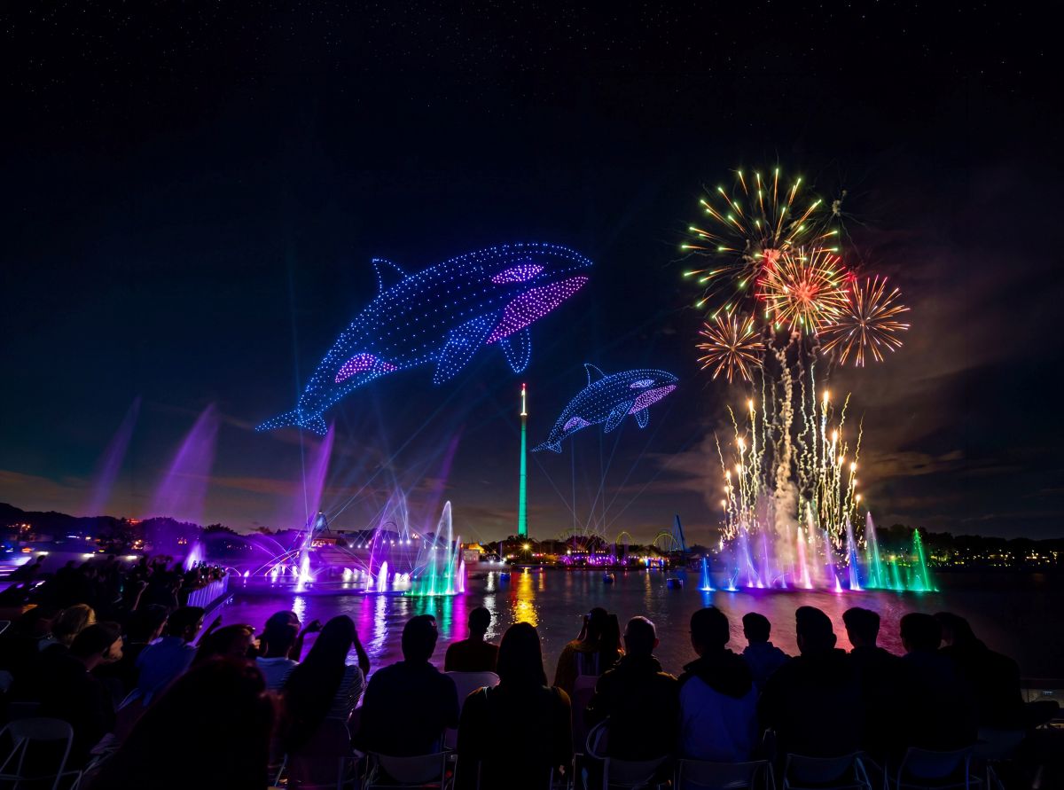 Nighttime waterfront show with glowing blue dolphin displays, colorful fountain jets, and fireworks over a seated crowd.