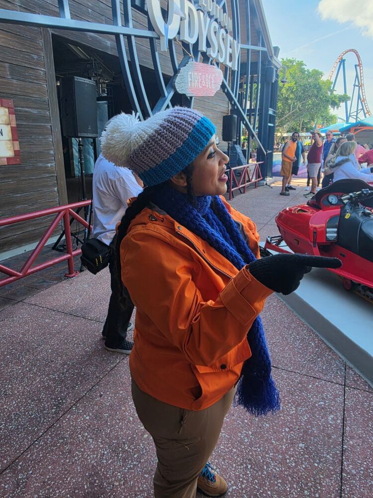 Woman in an orange jacket and blue knit scarf points toward a red amusement ride vehicle outside a theme park, with people in the background.