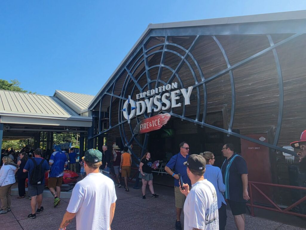 Crowd of people line up outside the Expedition Odyssey: Fire & Ice attraction at a theme park, a large circular metal framework above the entrance.