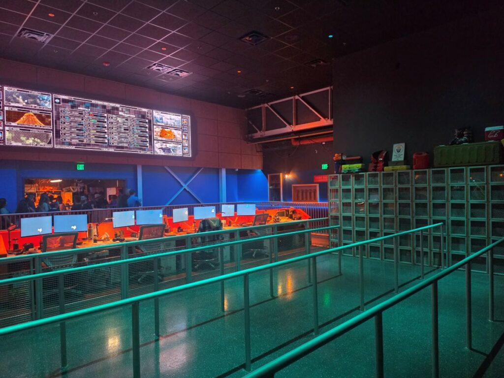Esports arena with blue and red lighting, row of computer workstations, and large wall screens showing stats and maps; railing in foreground.