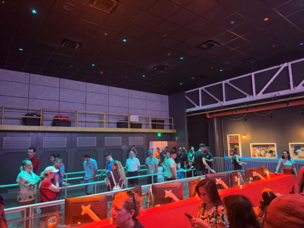 Busy indoor venue with people standing in line and walking along a mezzanine above red-lit workstations or counters.