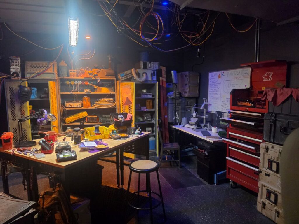 cluttered electronics workshop with shelves, tools, and a workbench filled with electronic parts and cables