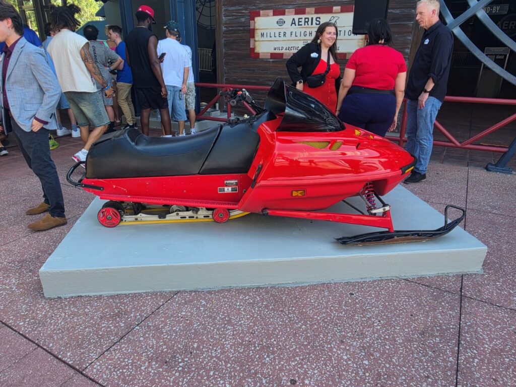 Red snowmobile on a gray raised display platform, with a crowd of people around it at an outdoor exhibit.