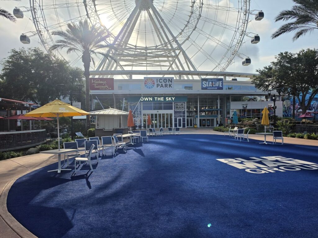 Outdoor area at Icon Park with a giant Ferris wheel in the background, blue-carpet seating area, and umbrellas.