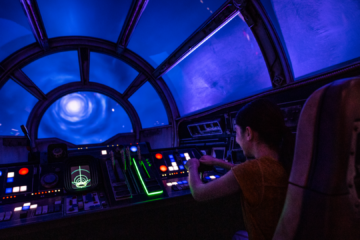 Person in an orange shirt sits at a sci‑fi cockpit with illuminated controls, blue space visible through a circular window.