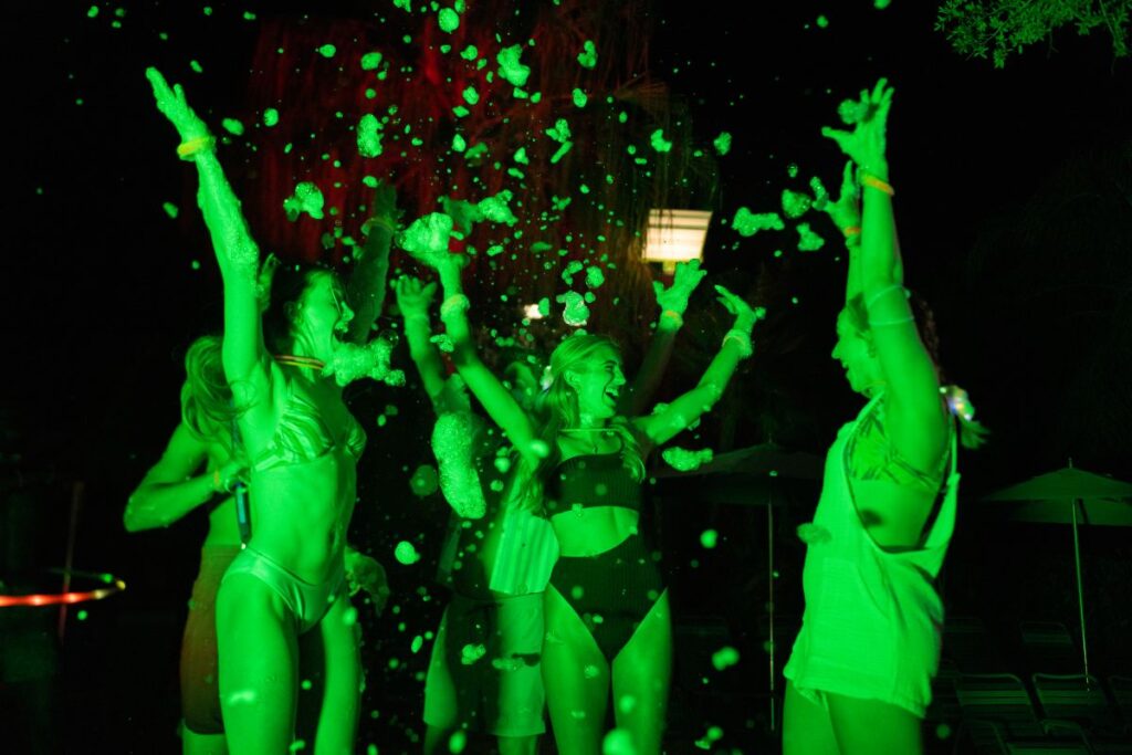 Group of friends dancing with foam in a club-like setting, neon green lighting, arms raised in celebration at night.