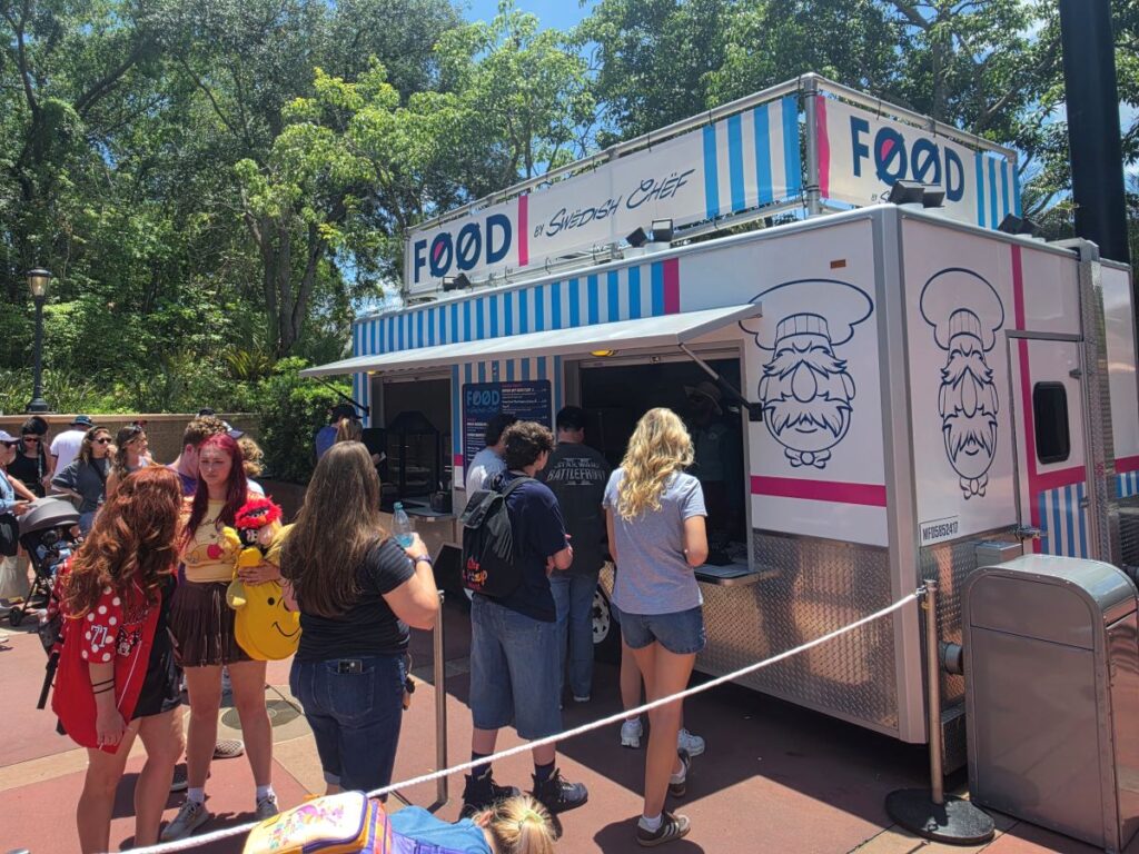Food truck with blue striped exterior serving customers; people line up outside on a sunny day.