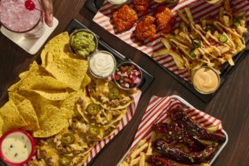 Top-down view of a shared spread: burgers, chicken wings, loaded nachos with jalapeños, fries, dips, and drinks on a dark wood table for Fridays United soccer menu
