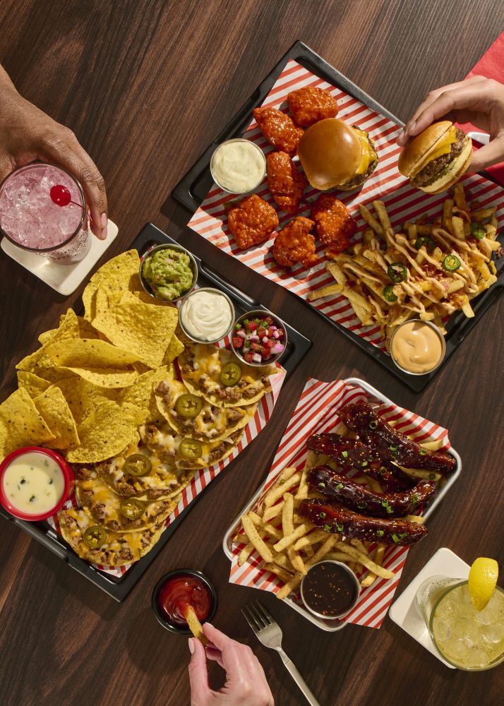 Top-down view of a shared spread: burgers, chicken wings, loaded nachos with jalapeños, fries, dips, and drinks on a dark wood table for Fridays United soccer menu