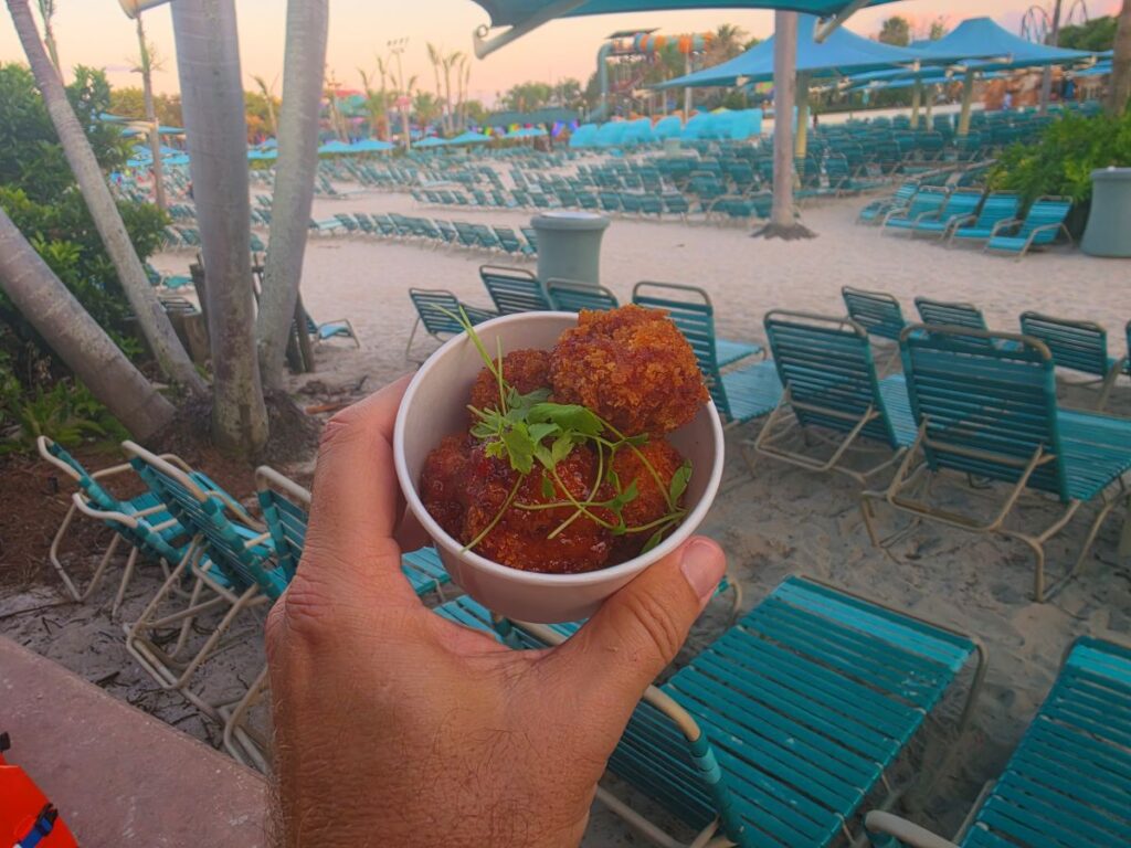 Hand holding a white bowl of fried croquettes with herbs at a beach resort with rows of turquoise lounge chairs and blue umbrellas.