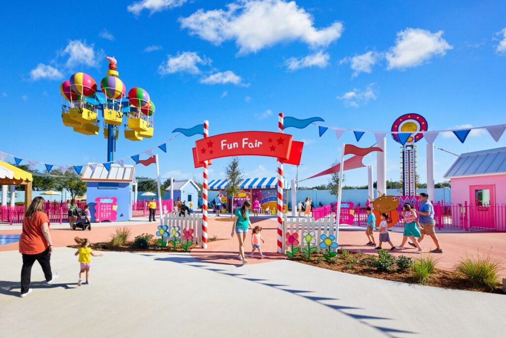Colorful fun fair entrance with a red striped arch reading 'Fun Fair' and families walking under a blue sky.