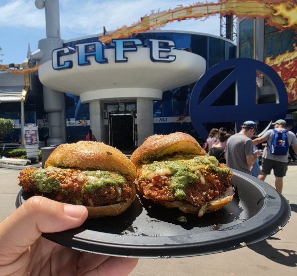 Two Garlic Parm Sliders with green topping on a plate held up outdoors, with people and a blue themed venue in the background.