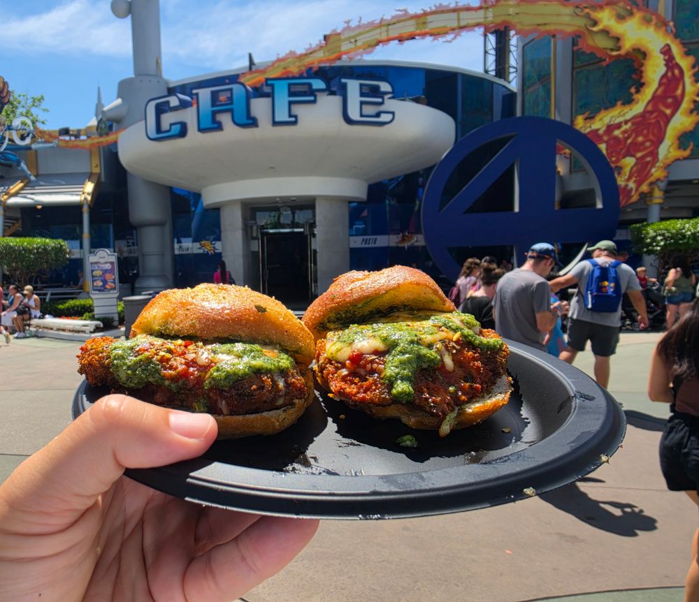 Two Garlic Park Sliders on a plate held up in front of a futuristic cafe entrance with a large blue circular logo and people walking by.