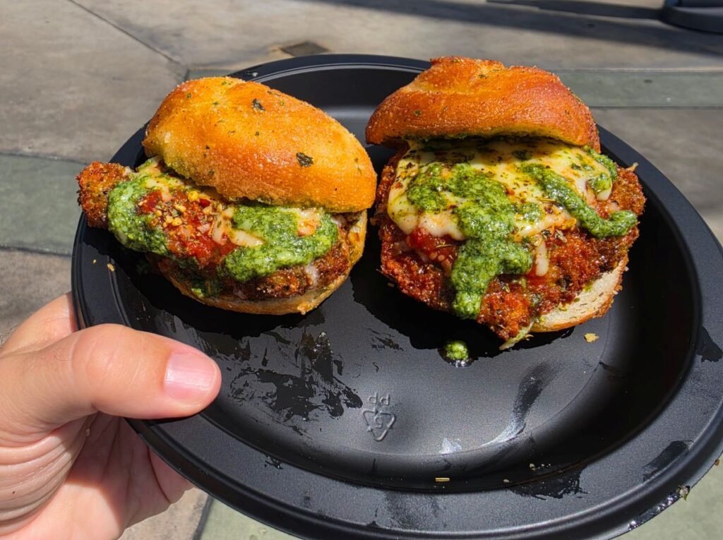 Two Garlic Parm Sliders on a black plate, with crispy patties, melted cheese, and green pesto sauce, held outdoors by a hand.