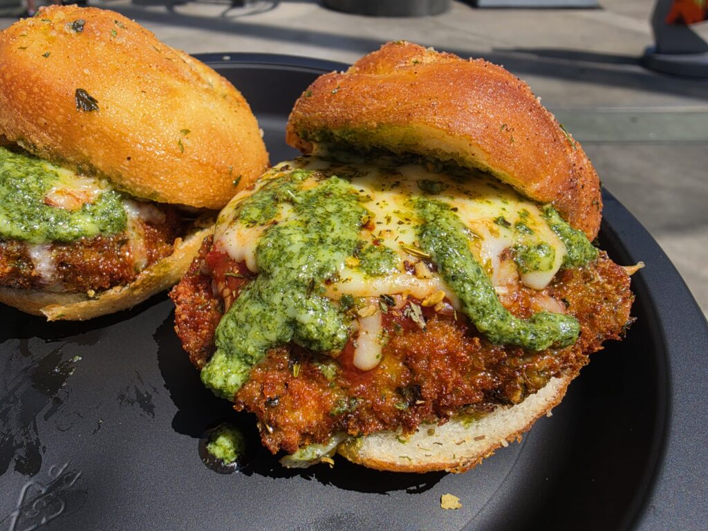 Two round, crispy-breaded buns with melted cheese and green herb pesto spilling over a dark plate.