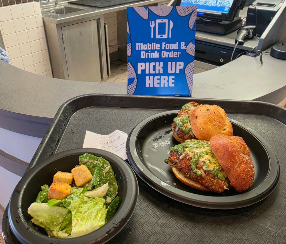 Blue sign reads 'Mobile Food & Drink Order Pick Up Here' at a cafe counter; on the tray are a side salad and Garlic Parm Sliders plate.