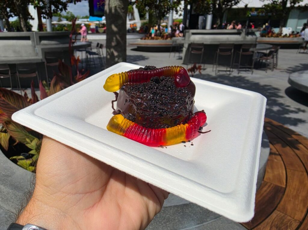 Chocolate dessert on a white disposable plate topped with gummy worms, served outdoors in a plaza setting for a Grab-N-Goof Epcot Review