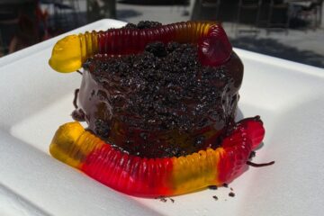 Chocolate cake with a glossy ganache, crushed cookie topping, surrounded by colorful gummy worms on a white plate for a Grab-N-Goof Epcot Review