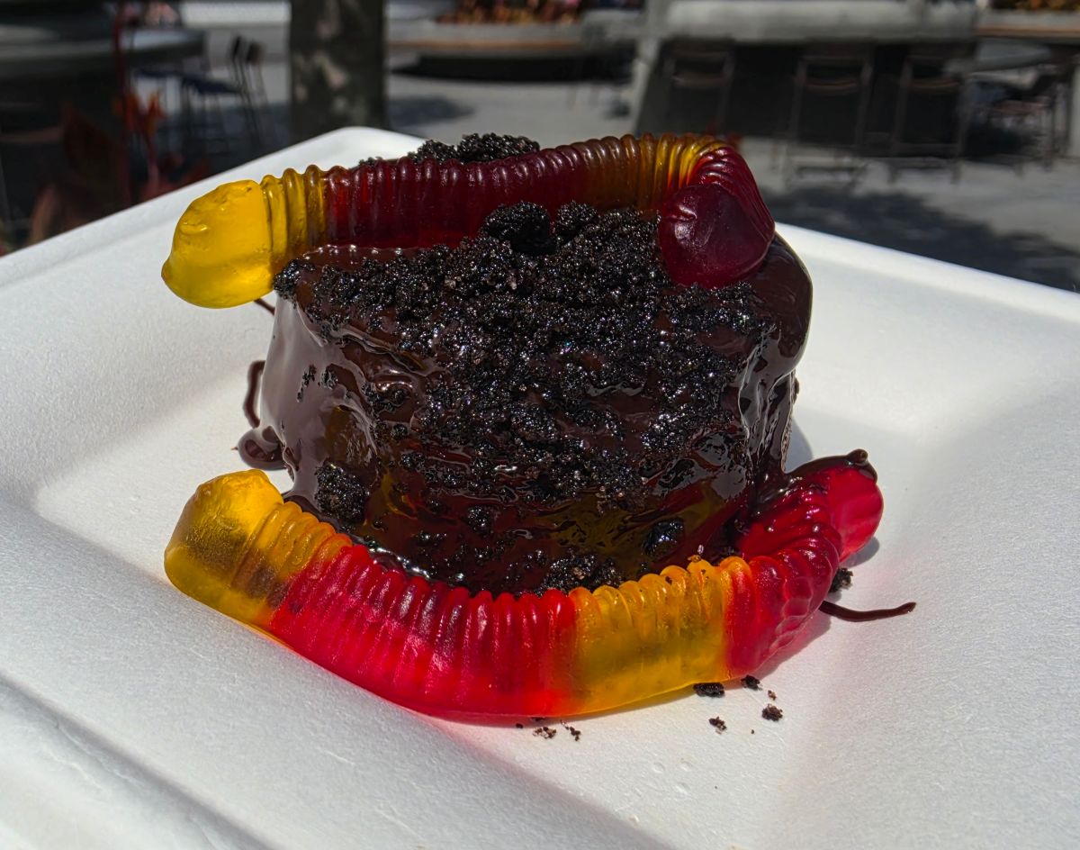 Chocolate cake with a glossy ganache, crushed cookie topping, surrounded by colorful gummy worms on a white plate for a Grab-N-Goof Epcot Review