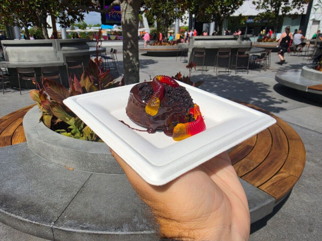 Chocolate cake on a white foam plate held up in an outdoor plaza with cafe tables and people in the background for a Grab-N-Goof Epcot review