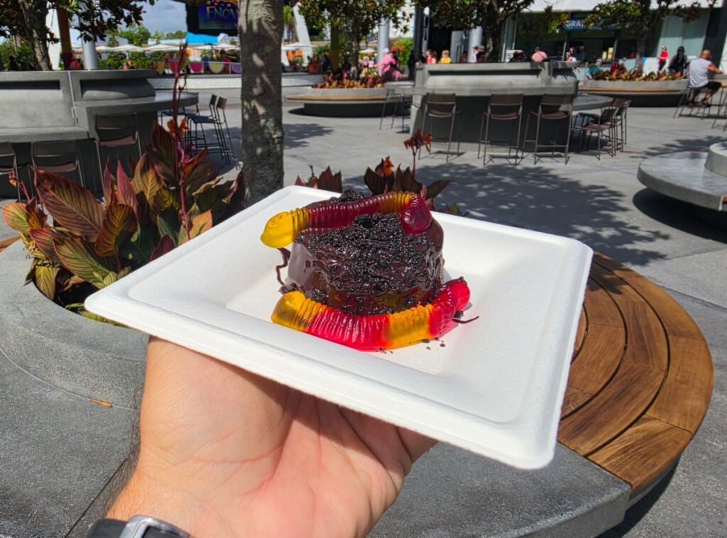 Person holds a white plate with a small chocolate cake adorned with gummy worms outdoors at a plaza or food court for a Grab-N-Goof Epcot review