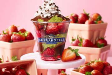 Dubai Style Chocolate Strawberry Ghirardelli dessert cup topped with whipped cream and chocolate drizzle, surrounded by fresh berries in pink bowls on a pink background.