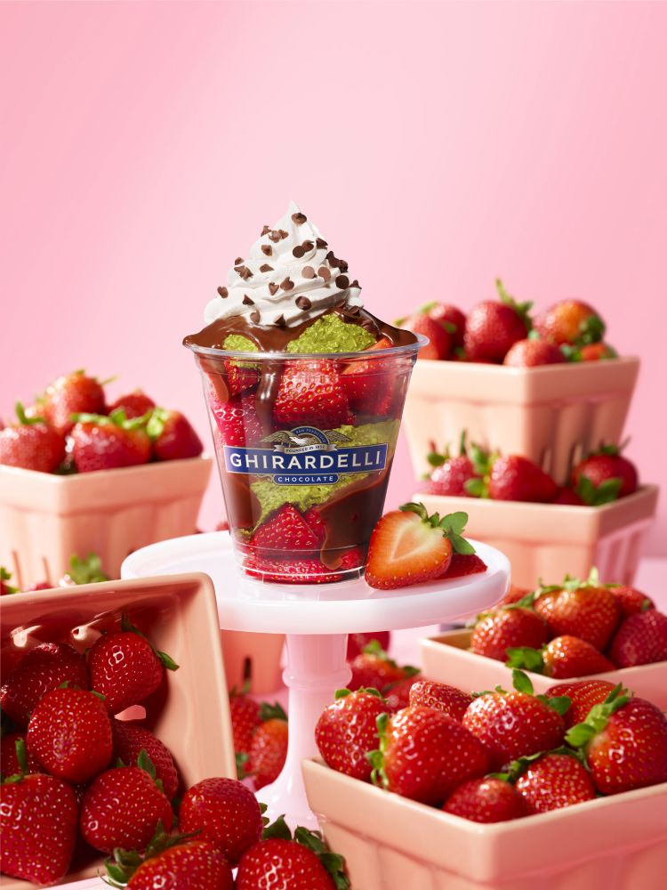 Dubai Style Chocolate Strawberry Ghirardelli dessert cup topped with whipped cream and chocolate drizzle, surrounded by fresh berries in pink bowls on a pink background.