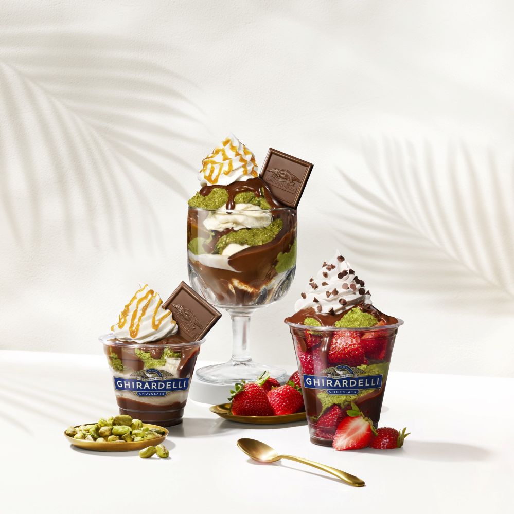 Ghirardelli chocolate sundaes with whipped cream, chocolate drizzle, and a square chocolate piece in a glass, surrounded by strawberries and pistachios