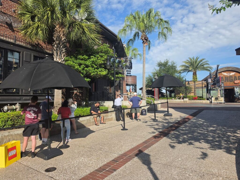People waiting in line behind a rope barrier with large black umbrellas along a brick-walled shopping area under a sunny blue sky.