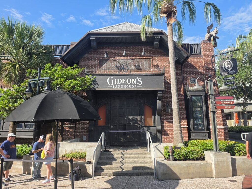 Brick bakehouse with a black sign reading 'Gideon's Bakehouse', flanked by palm trees and a large black umbrella in front, under a blue sky.