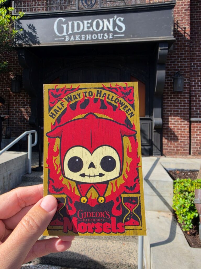 Promotional card with a red skeleton figure and 'Half Way to Halloween' text, held up in front of Gideon's Bakehouse sign.