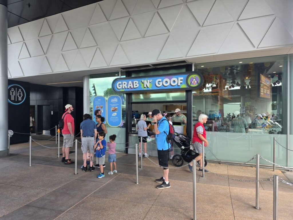 Exterior view of the Grab 'N' Goof food kiosk with a line of people waiting outside, including children and a stroller nearby.