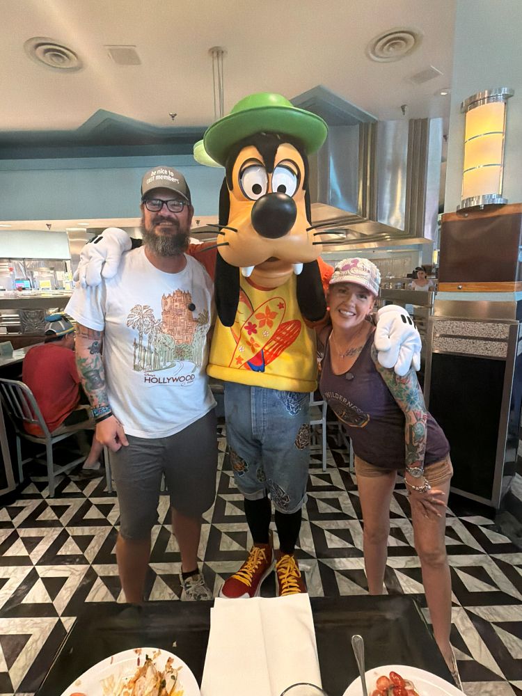 Man and woman pose with a Goofy character in a retro-style diner, smiling for the camera with plates on the table nearby.