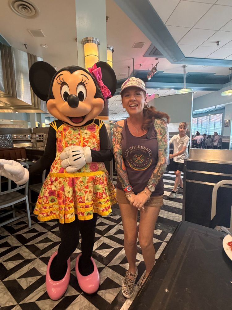 Minnie Mouse in a colorful floral dress stands beside a smiling woman with tattoos inside Hollywood & Vine Lunch.
