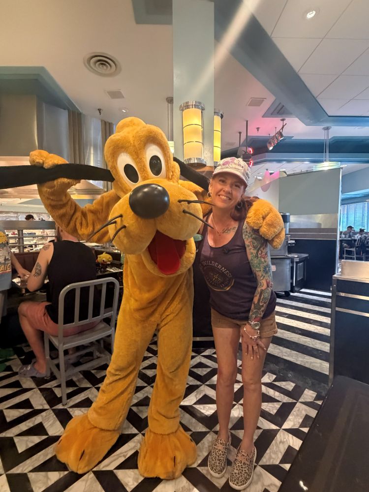 Woman with tattoos posing with a Pluto mascot in a retro diner interior.
