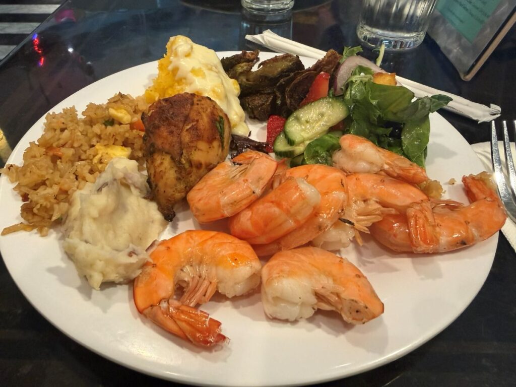 a Hollywood & Vine lunch plate with several large shrimp arranged over white rice, alongside mashed potatoes, fried plantains, and a green salad with cucumber slices and vegetables.
