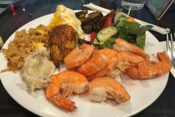 a Hollywood & Vine lunch plate with several large shrimp arranged over white rice, alongside mashed potatoes, fried plantains, and a green salad with cucumber slices and vegetables.
