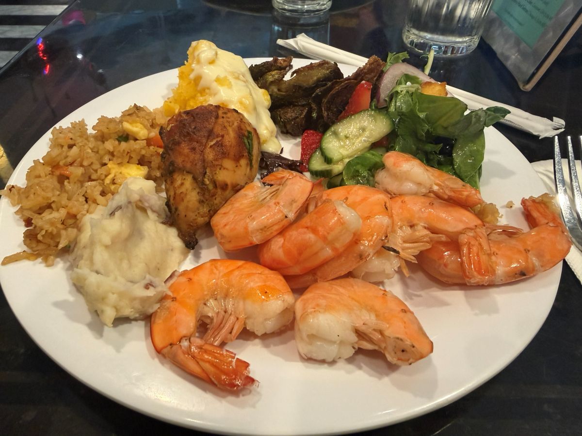 a Hollywood & Vine lunch plate with several large shrimp arranged over white rice, alongside mashed potatoes, fried plantains, and a green salad with cucumber slices and vegetables.