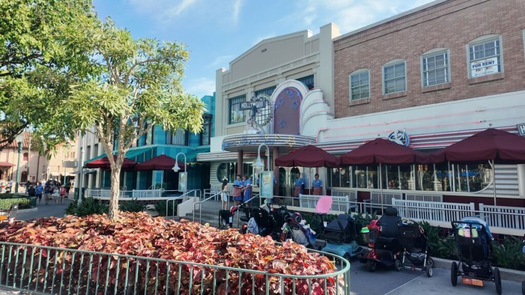 Sunlit street in a themed entertainment area with a vintage theater facade, red umbrellas, and parked strollers along the walkway.