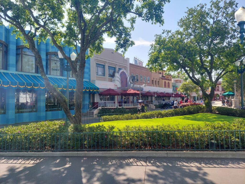 Colorful street view with blue striped storefronts, outdoor cafe seating under maroon umbrellas, and pedestrians along a sunny sidewalk. Trees and trimmed hedges line the green lawn in front of the row of shops.