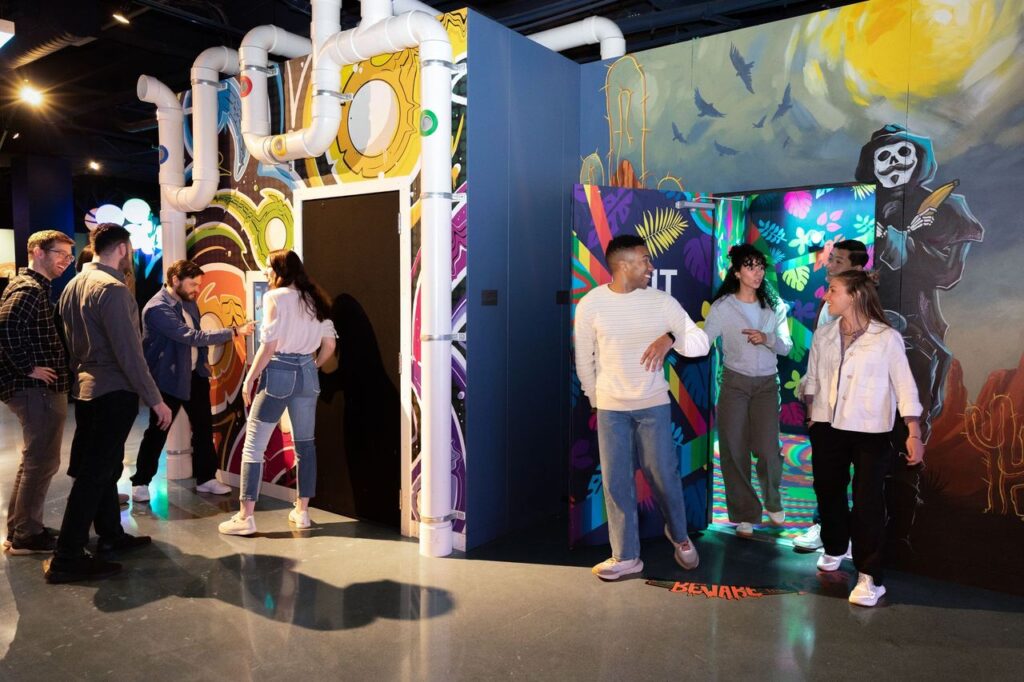 Group of people entering a black doorway in a bright, graffiti-covered interactive exhibit with colorful walls and piping structures nearby